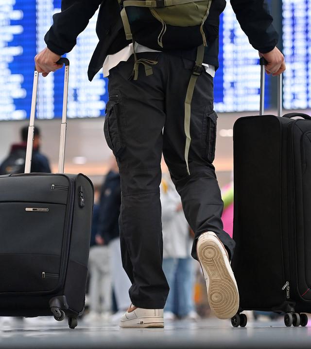 Passagier am Flughafen geht mit zwei Koffern auf Anzeigetafel zu.