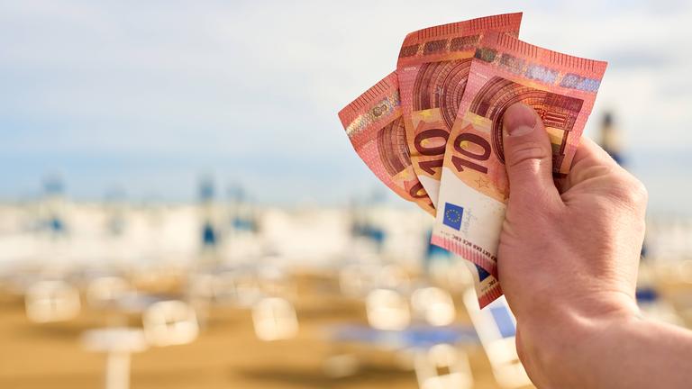 Ein Mann hält Euroscheine vor einer Reihe von Liegestühlen in Italien in der Hand.