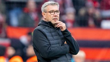 Fußballtrainer Urs Fischer trägt eine schwarze Weste und blickt nachdenklich von der Seitenlinie aufs Spielfeld.