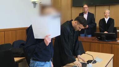 Das Bild zeigt einen Gerichtsaal. Im Vordergrund der Angeklagte, der mit einer Mappe sein Gesicht verdeckt. 