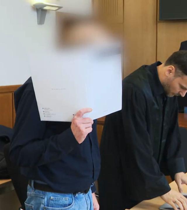 Das Bild zeigt einen Gerichtsaal. Im Vordergrund der Angeklagte, der mit einer Mappe sein Gesicht verdeckt. 