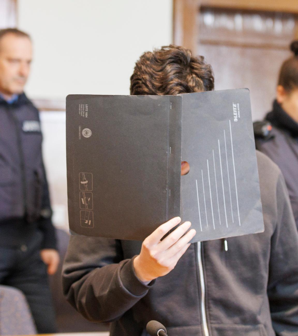 Der Angeklagte ist im Gerichtssaal zu sehen, er verdeckt sein Gesicht. Im Hintergrund sind zwei Polizist*innen zu sehen.