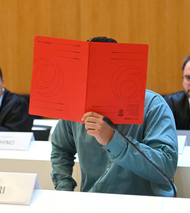 Der Angeklagte im Verhandlungssaal, verbirgt sein Gesicht hinter einem Schnellhefter. Hinten seine Anwälte und Verteidiger.