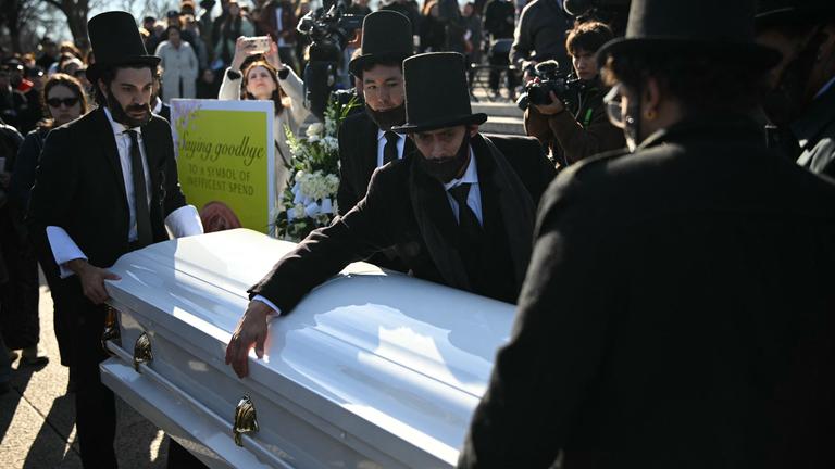 Männer, gekleidet wie der ehemalige US-Präsident Abraham Lincoln, öffnen einen Sarg, in den die Menschen während einer „Beerdigung“ für den Penny am Lincoln Memorial am 20. Dezember 2025 Pennys werfen können, um ihre Ehrerbietung zu erweisen.