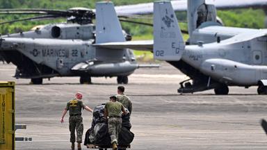 Militräflugzeuge und -hubschrauber und drei Soldaten, die etwas über das Rollfeld schieben.