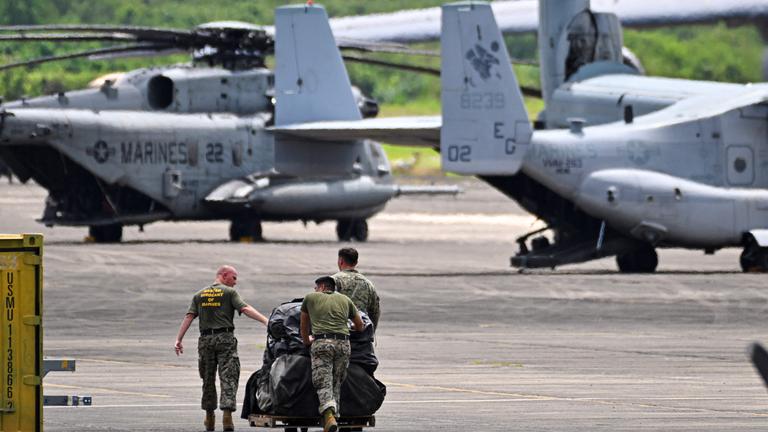 Militräflugzeuge und -hubschrauber und drei Soldaten, die etwas über das Rollfeld schieben.