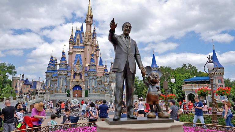 Eine Statue von Walt Disney, der Micky Maus an der Hand hält, steht in einem Vergnügungspark. Dahinter laufen dutzende Touristen vor einem Märchenschloss.