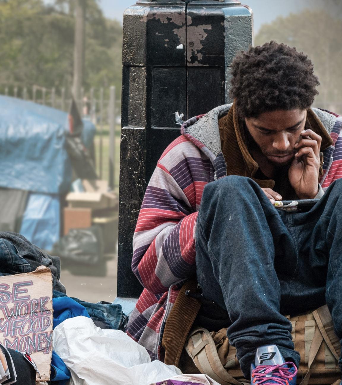 USA extrem: Obdachlos in Los Angeles": in der linken Bildhälfte improvisiertes Zelt aus Planen mit US-Fahne vor einem Zaun vor parkähnlichem Grundstück im Hintergrund, davor im Vordergrund Müllsäcke und Pappschild mit Schriftzug PLEASE HELP I WORK FOR ROOM FOOD MONEY und in der rechten Bildhälfte ein junger Mann, der auf einer Tasche sitzt, am unteren Teil einer Straßenlaterne lehnt und auf sein Smartphone schaut.