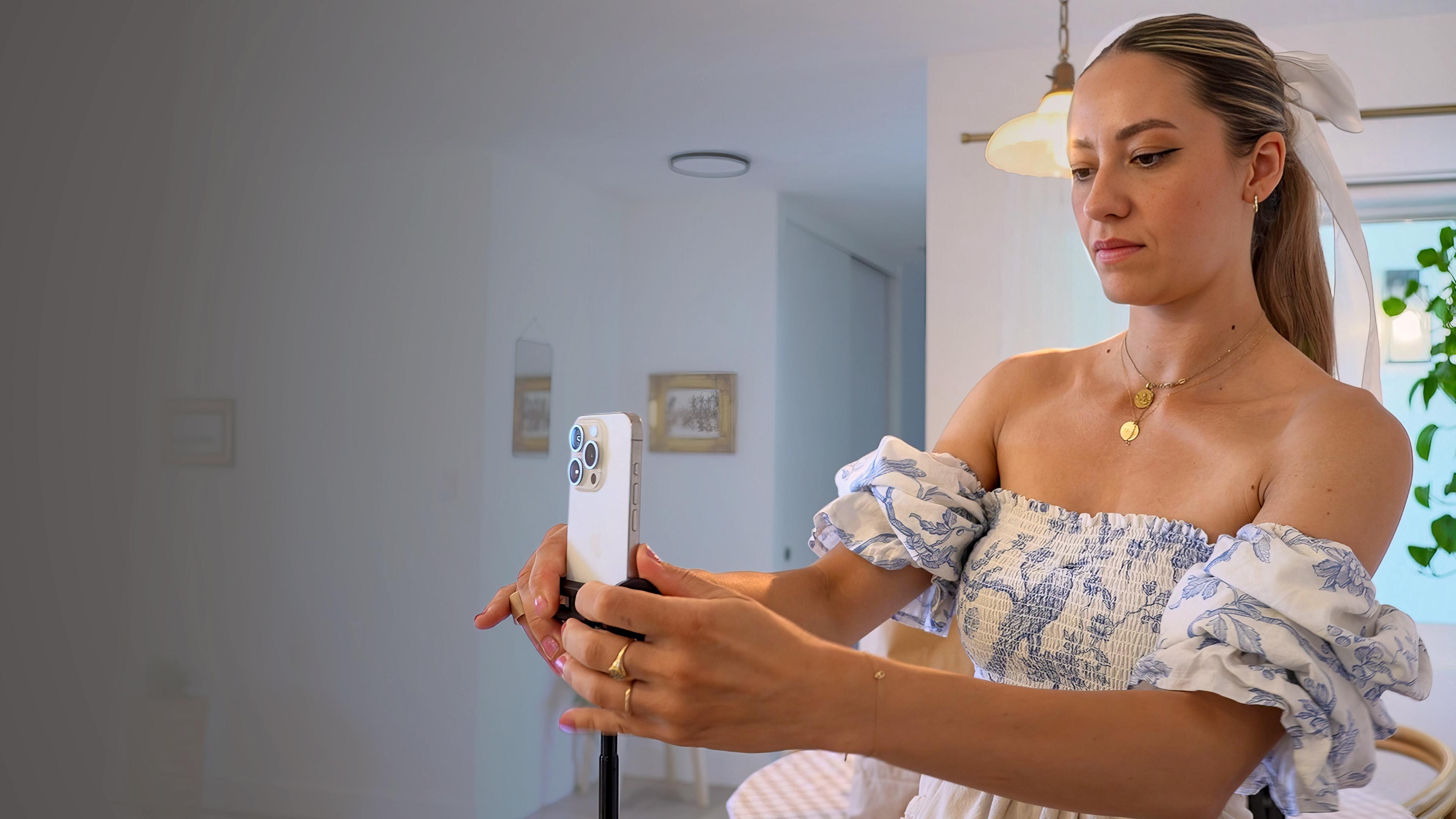 Influencerin Alexia Delarosa richtet ihr Handy auf einem Stativ auf sich selbst aus. Sie trägt ein schulterfreies Kleid und steht in häuslicher Umgebung in einem hellen Raum.