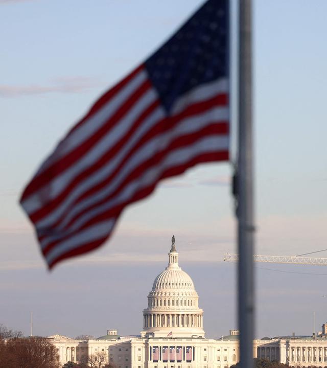 Die US-amerikanische Flagge weht im Vordergrund, weiter hinten ist das Kapitol in Washington zu sehen.