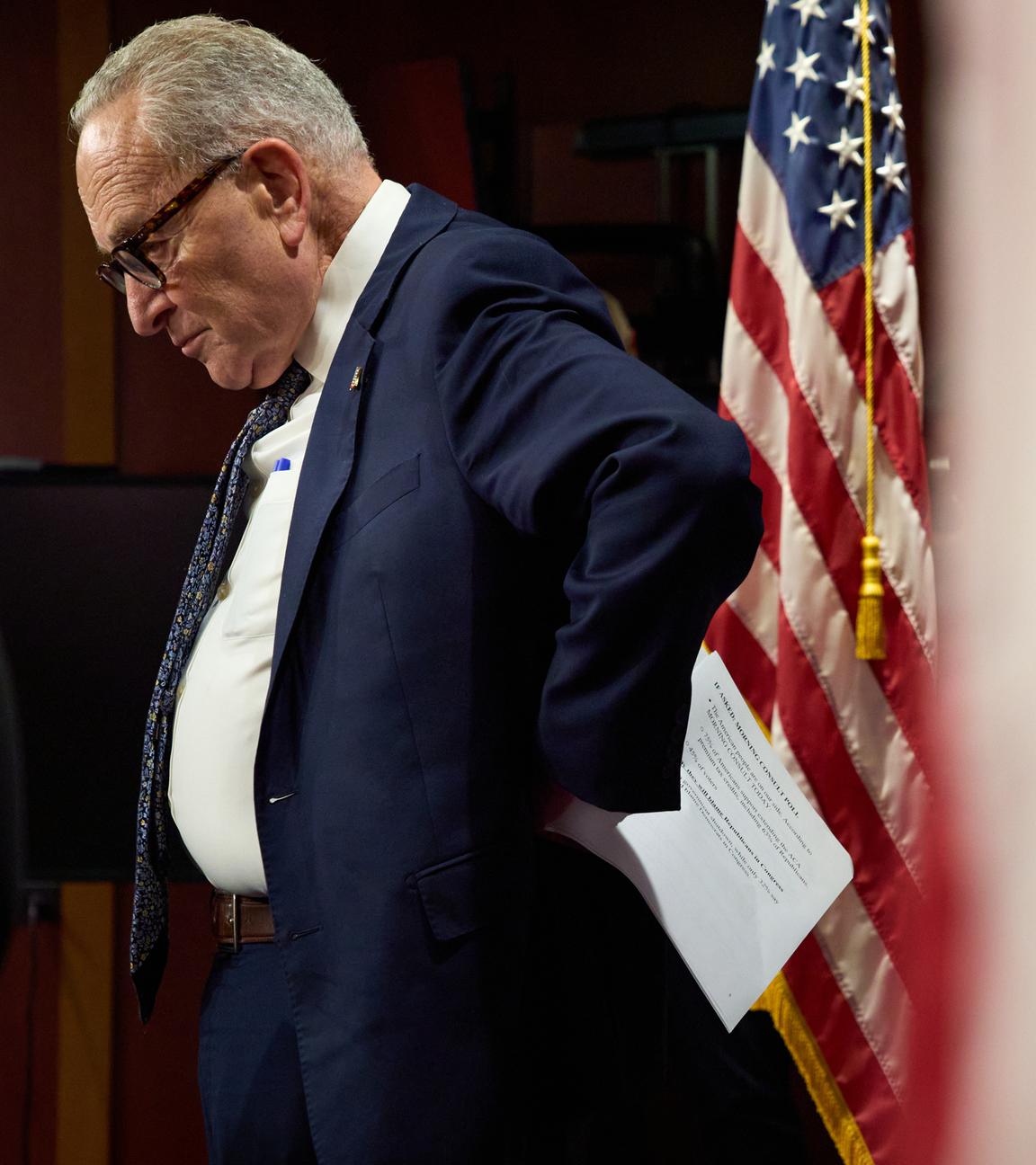 Minderheitsführer des US-Senats Charles "Chuck" Schumer ist zu sehen wie er seitlich steht und auf den Boden schaut. Neben ihm hängt eine amerikanische Flage