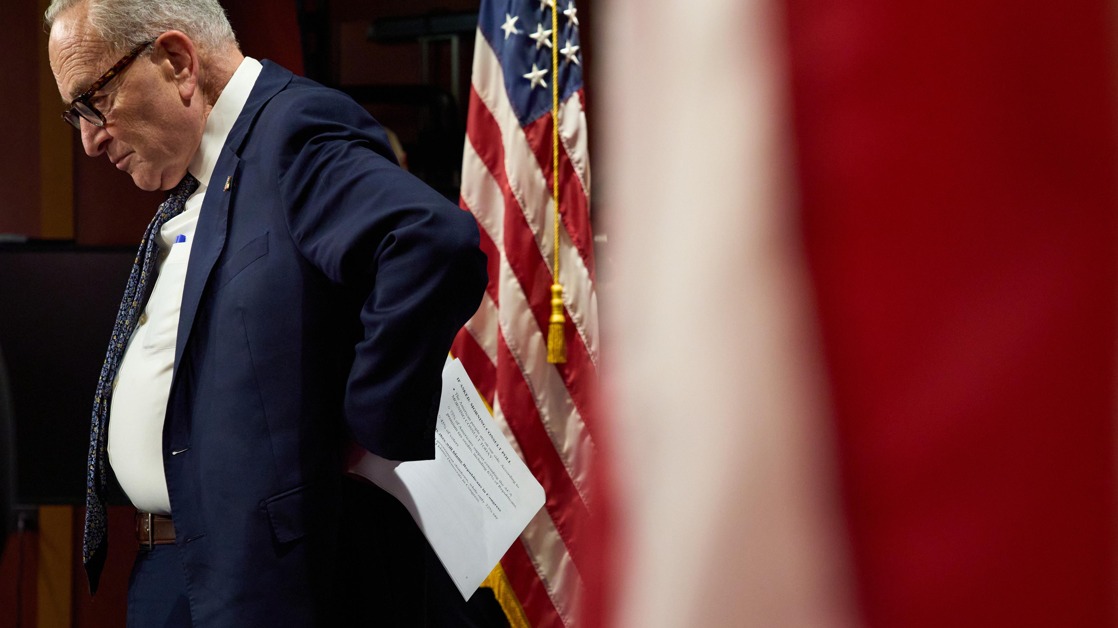 Minderheitsführer des US-Senats Charles "Chuck" Schumer ist zu sehen wie er seitlich steht und auf den Boden schaut. Neben ihm hängt eine amerikanische Flage