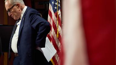 Minderheitsführer des US-Senats Charles "Chuck" Schumer ist zu sehen wie er seitlich steht und auf den Boden schaut. Neben ihm hängt eine amerikanische Flage
