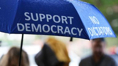 27.10.2026, USA, Coral Gables: Eine Person trägt vor einem Wahllokal einen Regenschirm mit der Aufschrift "Support Democracy" und "No Dictators" während die vorzeitige Stimmabgabe für die Zwischenwahlen 2026 stattfindet.