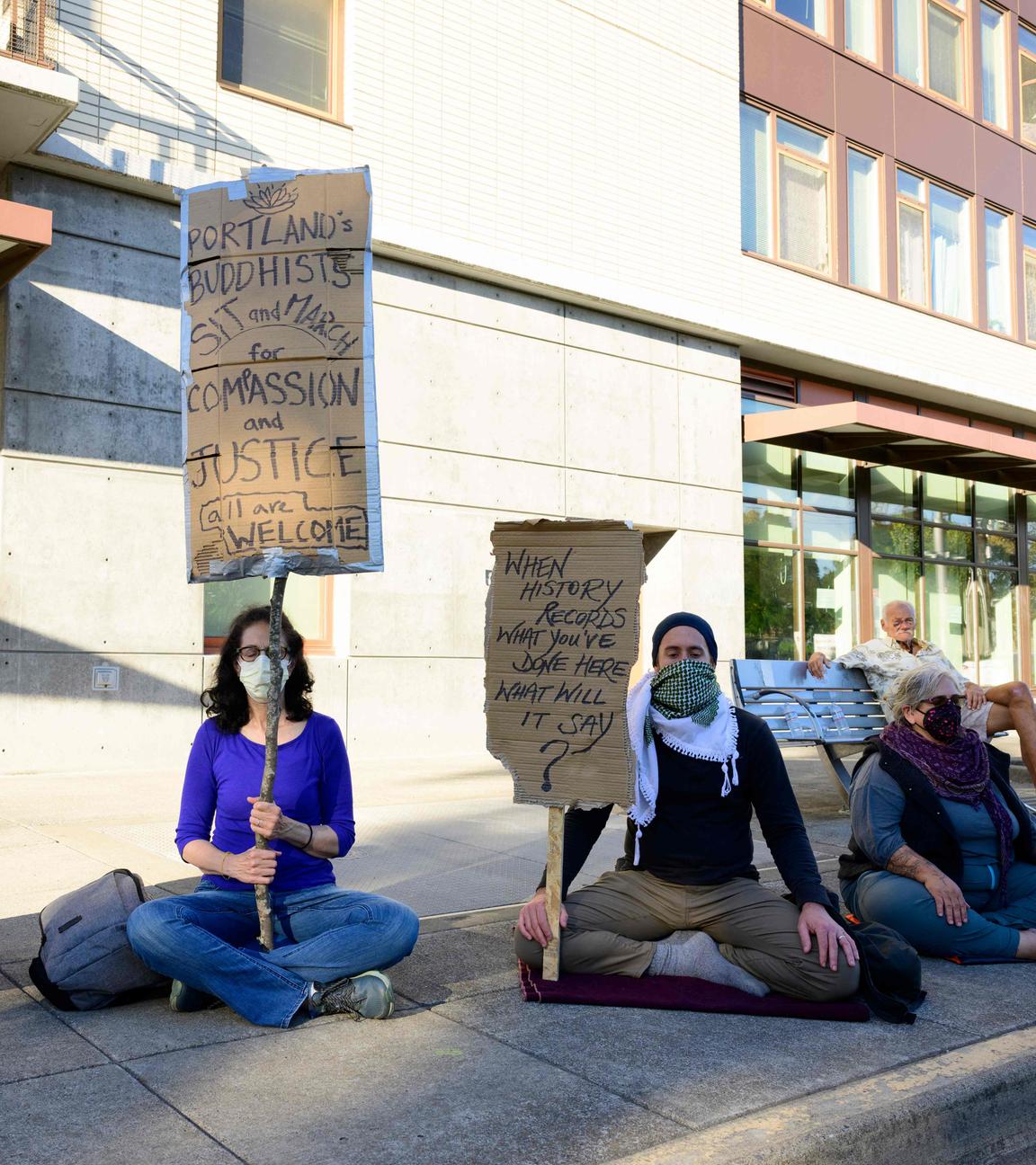Mehrere Demosntranten sitzen in Portland, USA vor einem Gebäude der Migrationsbehörde mit Schildern in der Hand auf der Straße.