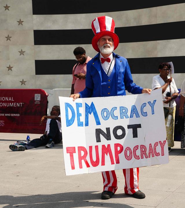 Ein Mann ist gekleidet in den Farben der USA und hält ein Demonstrationsplakat in den Händen.