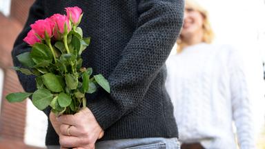 Ein Mann hält einen Strauß Rosen hinter seinem Rücken während eine Frau vor ihm steht.