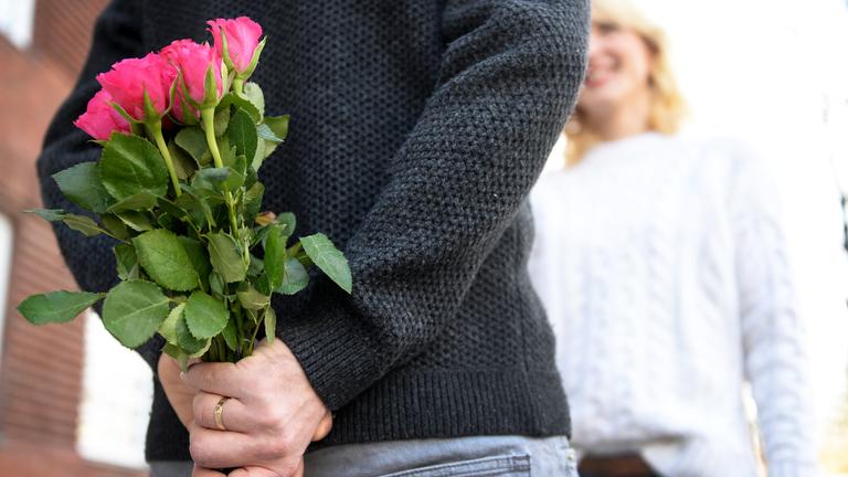 Ein Mann hält einen Strauß Rosen hinter seinem Rücken während eine Frau vor ihm steht.