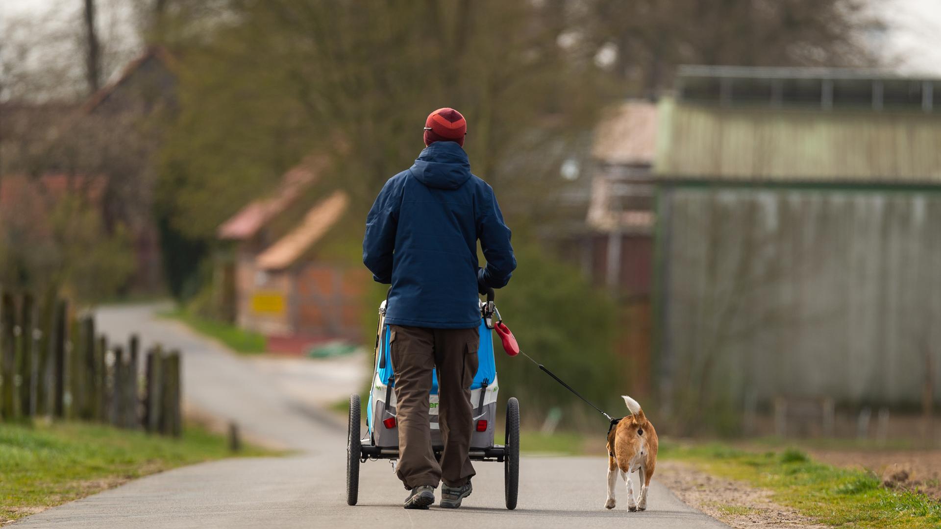 Ein Vater geht mit Hund und Kinderwagen draußen auf einem Weg spazieren.