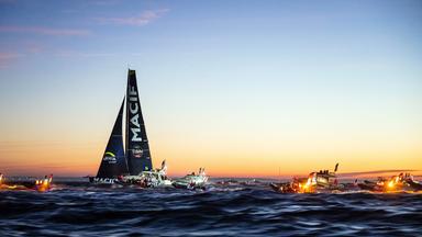 Ein Segelschiff auf dem Meer umgeben von kleineren Motorbooten im Sonnenuntergang.