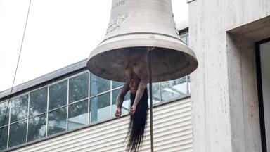 Eine nackte tätowierte Frau mit langen Haaren hängt wie ein Schlägel in einer Glocke.