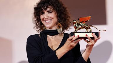 Eine Frau mit schulterlangen braunen Locken und im schwarzen Abendkleid präsentiert lächelnd eine goldene Trophäe, die einen geflügelten Löwen darstellt.