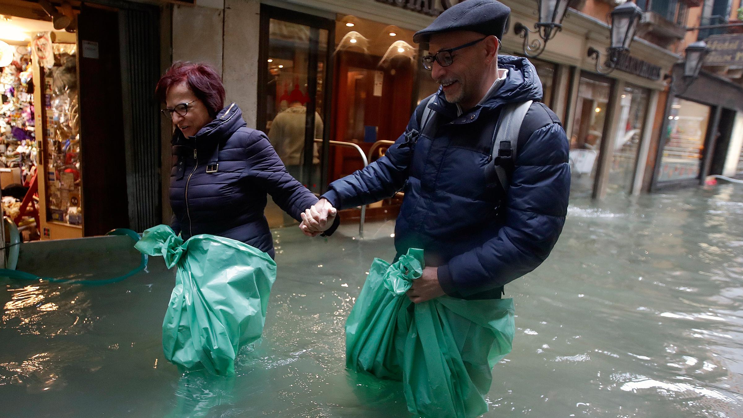 Ein Paar watet knietief in grünen Müllsäcken im Hochwasser einer überfluteten Straße. Am 15.11.2019 in Venedig, Italien.