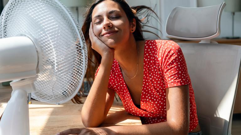 Lächelnde Frau sitzt mit elektrischem Ventilator am Schreibtisch.