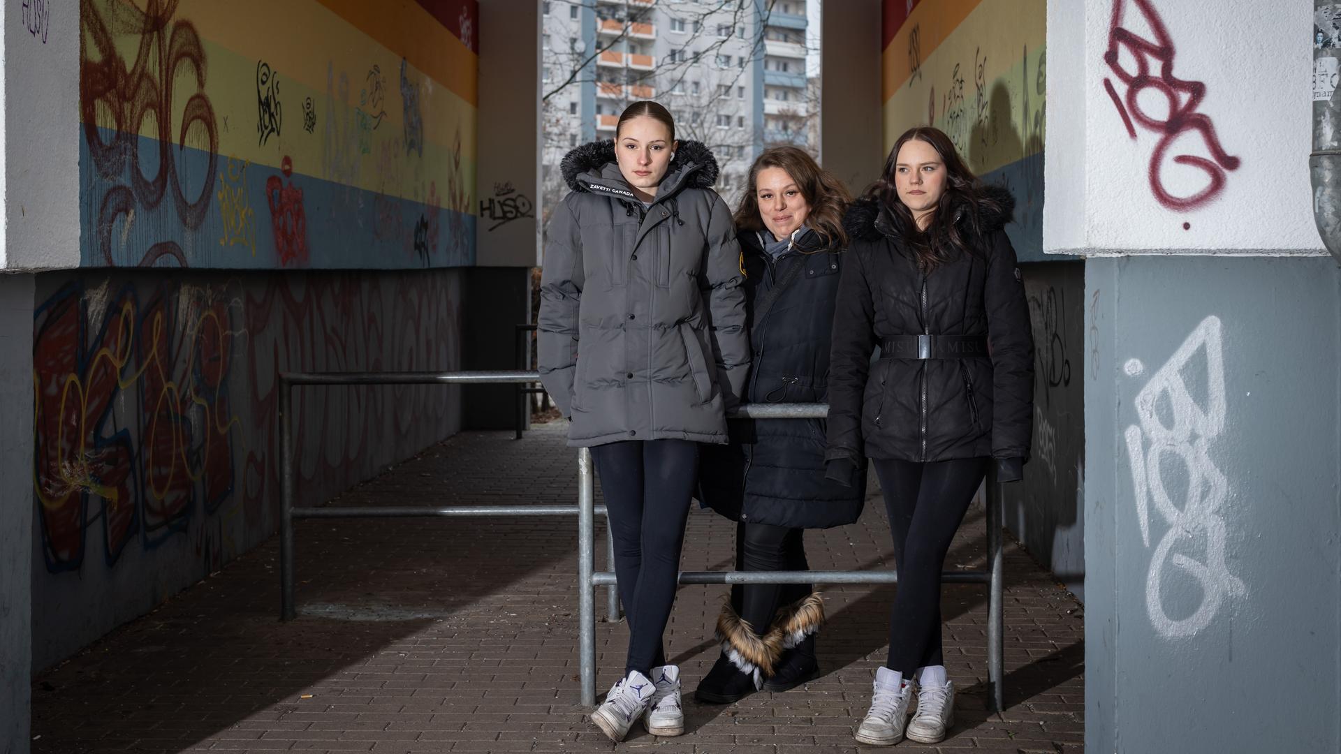 Angie (18), Luisa (16) und ihre Mutter Silvana (36) stehen an Stangen gelehnt in einem Hausdurchgang. In dem Gang befinden sich Graffitis, dahinter steht ein Hochhaus.