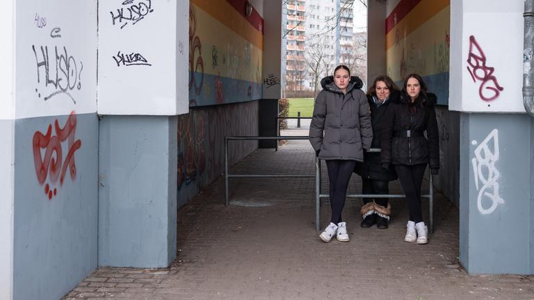 Angie (18), Luisa (16) und ihre Mutter Silvana (36) stehen an Stangen gelehnt in einem Hausdurchgang. In dem Gang befinden sich Graffitis, dahinter steht ein Hochhaus.
