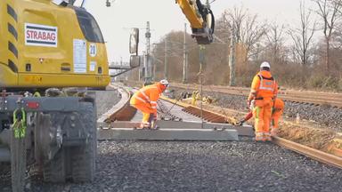 Das Bild zeigt: Mehrere Personen arbeiten auf einer Baustelle an Bahngleisen. Ein Bagger hebt Gleiselemente an, während die Arbeiten an der Strecke fortgesetzt werden.