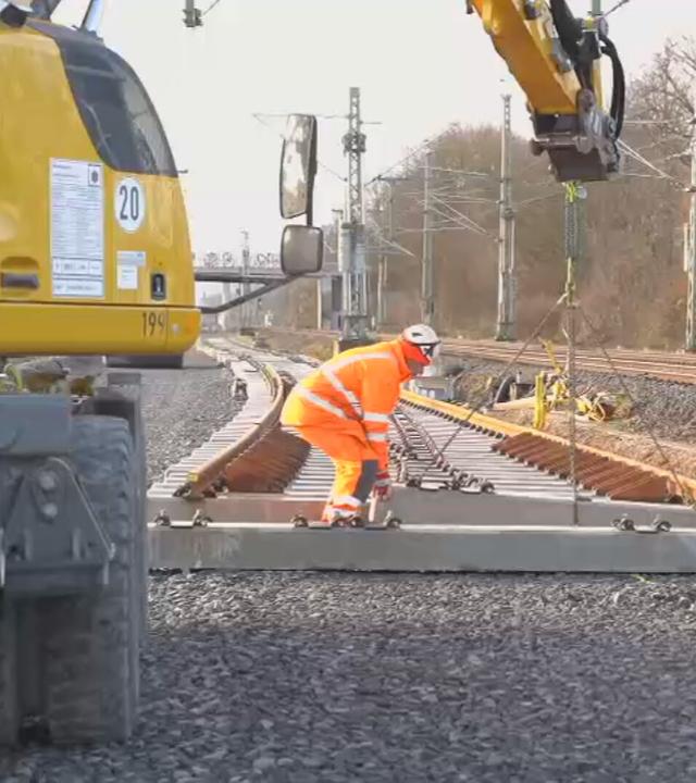 Das Bild zeigt: Mehrere Personen arbeiten auf einer Baustelle an Bahngleisen. Ein Bagger hebt Gleiselemente an, während die Arbeiten an der Strecke fortgesetzt werden.