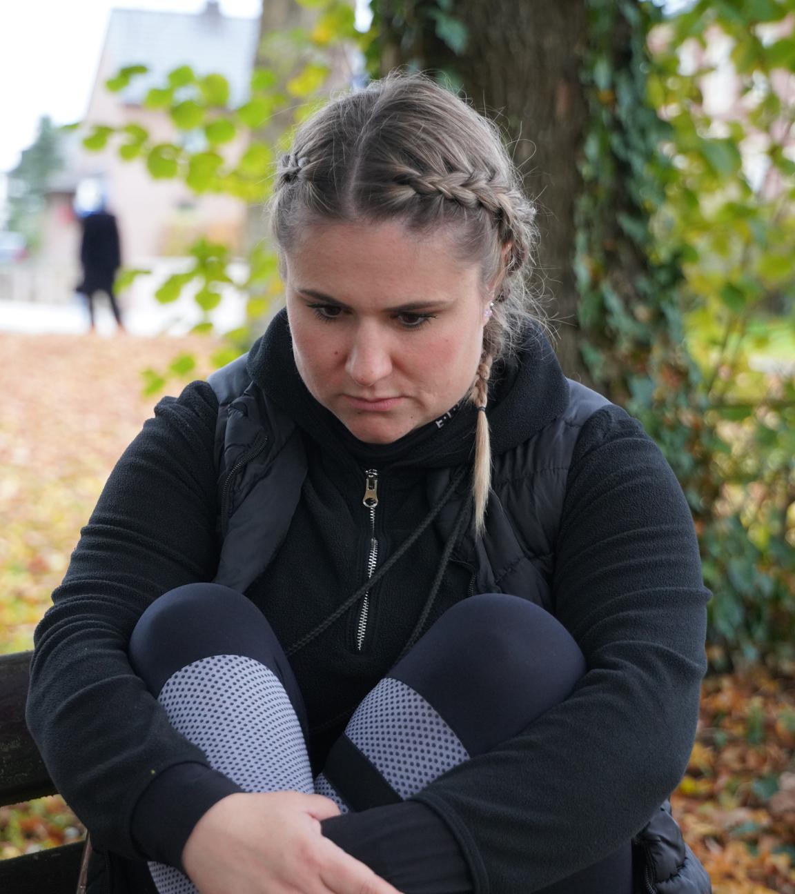 Ann-Kristin sitzt auf einer Bank, den Blick nach unten gerichtet. Sie umfasst ihre Beine mit den Händen. Im Hintergrund ist ein Mann in schwarz zu sehen. 
