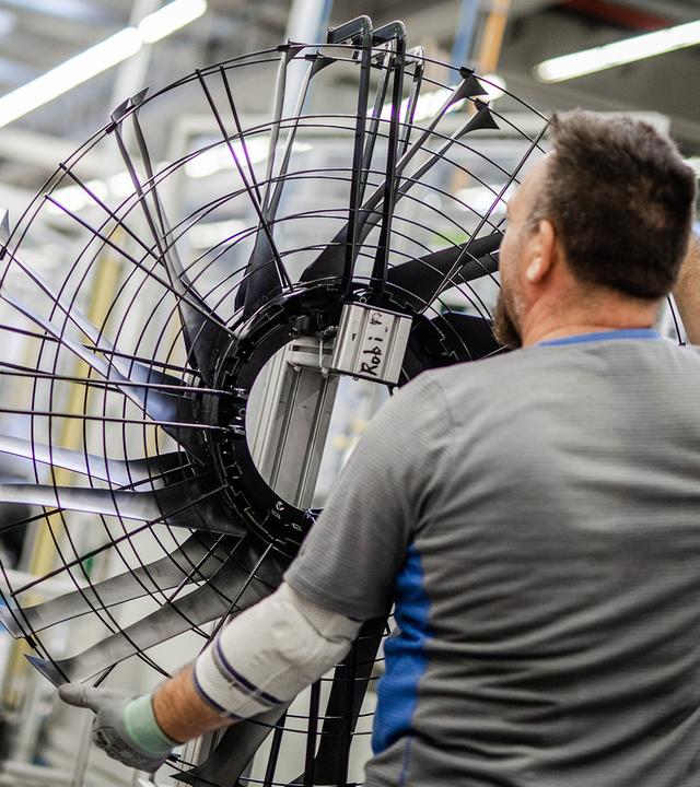 Arbeiter von hinten mit großem Ventilator-Rad in der Hand. Daneben Kartons von EBM-Papst