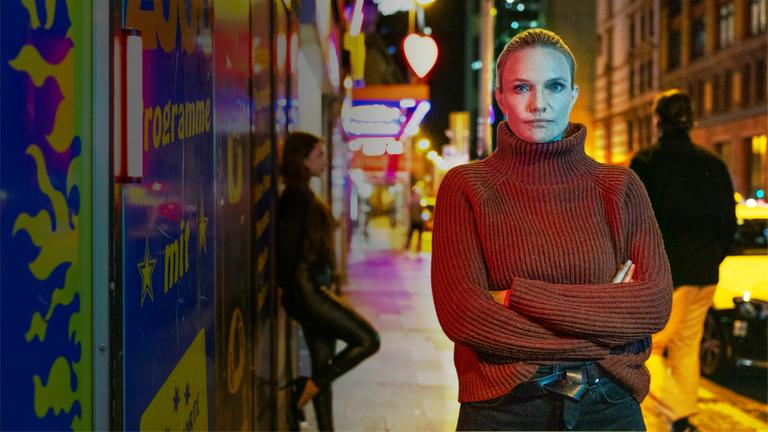 Sarah Tacke steht vor der Kulisse eines Rotlichtviertels. Im Hintergrund lehnt eine Frau in Lederoutfit an einer Wand, ein Mann läuft die Straße entlang.