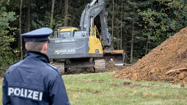 Ein von hinten fotografierter Polizist nimmt einen großen Teil der linken Bildhälfte ein. Auf seiner dunkelblauen Jacke steht in großen weißen Buchstaben „Polizei“. Sein Kopf ist einem Bagger zugewandt, der dahinter vor Bäumen steht.