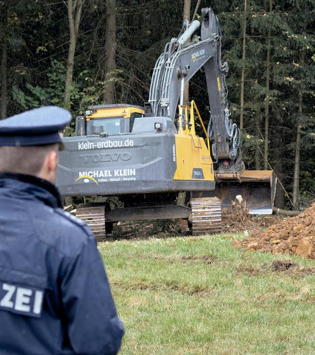 Ein von hinten fotografierter Polizist nimmt einen großen Teil der linken Bildhälfte ein. Auf seiner dunkelblauen Jacke steht in großen weißen Buchstaben „Polizei“. Sein Kopf ist einem Bagger zugewandt, der dahinter vor Bäumen steht.