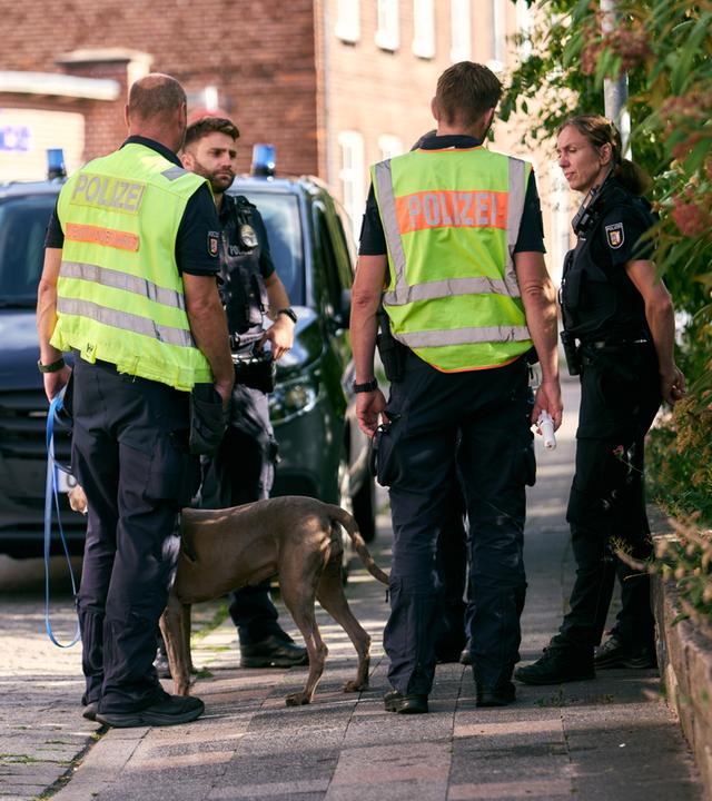 Mehrere Polizeikräfte stehen mit einem Hund neben einem Polizeifahrzeug auf einem Gehweg in einem Wohngebiet.