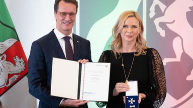 Auf dem Bild ist auf der linken Seite die Schauspielerin Veronica Ferres mit ihrer Auszeichnung in der Hand zu sehen. Auf der rechten Seite ist ein Mann zu sehen, der eine Urkunde in der Hand hält.