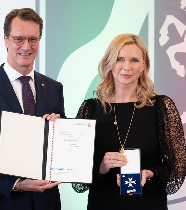 Auf dem Bild ist auf der linken Seite die Schauspielerin Veronica Ferres mit ihrer Auszeichnung in der Hand zu sehen. Auf der rechten Seite ist ein Mann zu sehen, der eine Urkunde in der Hand hält.