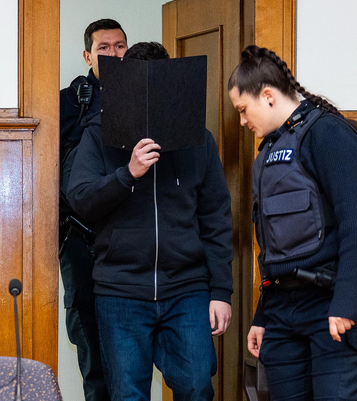 Der Angeklagte wird von Justizvollzugsbeamten einen Sitzungssaal des Landgericht Saarbrücken geführt und hält beim Betreten eine schwarze Akte vor sein Gesicht.