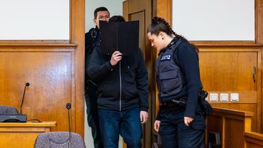 Der Angeklagte wird von Justizvollzugsbeamten einen Sitzungssaal des Landgericht Saarbrücken geführt und hält beim Betreten eine schwarze Akte vor sein Gesicht.