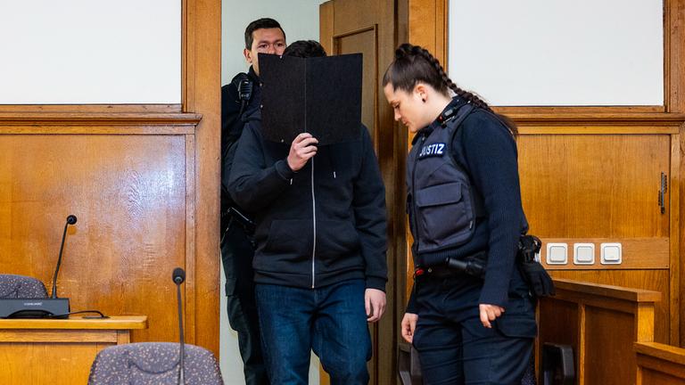 Der Angeklagte wird von Justizvollzugsbeamten einen Sitzungssaal des Landgericht Saarbrücken geführt und hält beim Betreten eine schwarze Akte vor sein Gesicht.