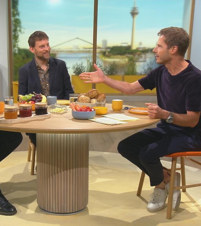 Wilson Gonzalez Ochsenknecht, Alexander Finkenwirth und Florian Weiss sitzen im Gespräch vertieft an einem runden Tisch. Der Tisch ist als Frühstückstisch reich gedeckt.