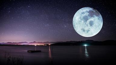 Eine Meeresküste bei einer Vollmondnacht. Der Himmel ist voller Sterne, im Hintergrund sieht man Berge. Der Mond ist riesig und sehr deutlich zu sehen.