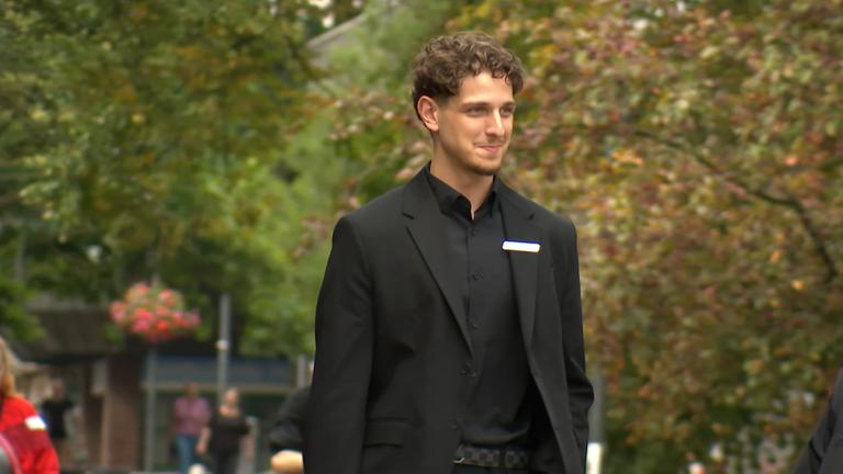 Ein Mann geht in ein Gebäude. Er trägt einen Anzug und streckt den Arm aus, um die Tür aufzumachen.