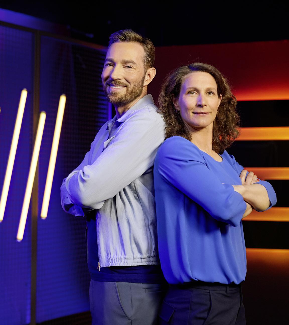 Moderator Sven Voss und Expertin Kathrin Lehmann stehen mit verschränkten Armen Rücken an Rücken vor LED-Leuchtröhren und blicken in die Kamera.