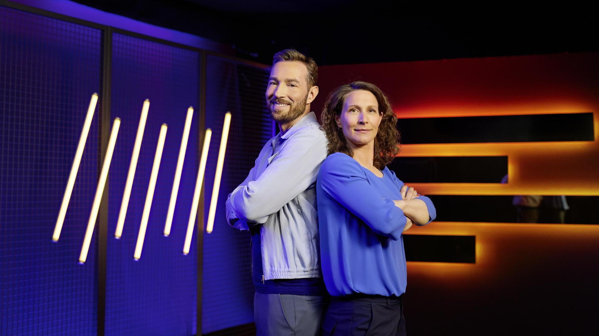 Moderator Sven Voss und Expertin Kathrin Lehmann stehen mit verschränkten Armen Rücken an Rücken vor LED-Leuchtröhren und blicken in die Kamera.