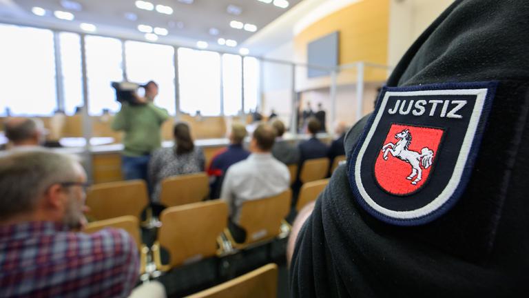 Ausschnitt aus dem Gerichtssaal. Es sind einige Besucher anwesend. Außerdem ist Personal der Justiz zu sehen.