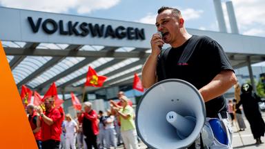 Vor einem Gebäude mit dem großen Schriftzug „Volkswagen“ spricht ein Mann in ein Megafon. Hinter ihm stehen mehrere Menschen mit roten Fahnen, einige applaudieren. Die Aufnahme zeigt eine Protestaktion bei Volkswagen an einem sonnigen Tag.